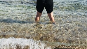 Una persona de pie en agua poco profunda, con los brazos levantados, usando un sombrero y un conjunto negro.