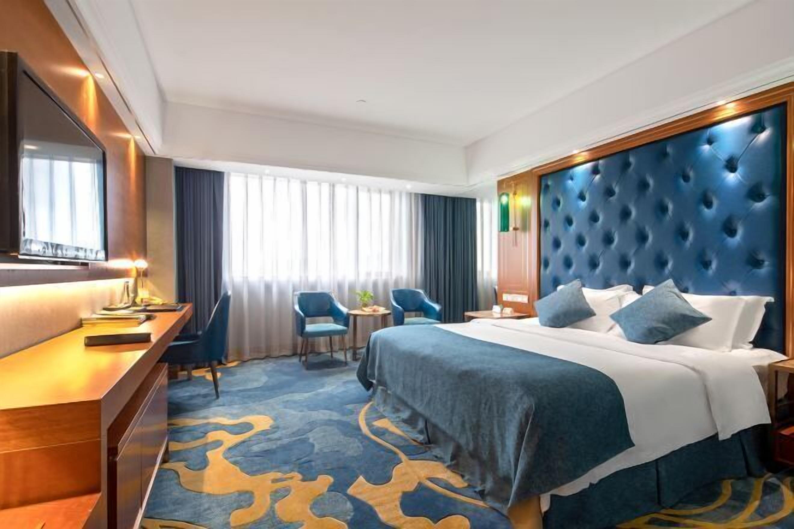 A hotel room with a large bed, a desk, two chairs, a TV, and a window with curtains.