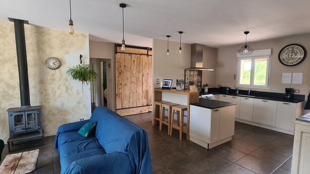 Una cocina moderna con una isla central, armarios de madera y una estufa. En la sala de estar, hay un sofá azul y una mesa de madera.