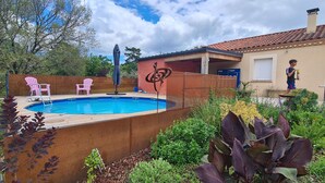 Un patio trasero con piscina, un porche cubierto y un jardín con diversas plantas y flores.