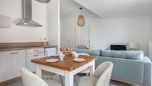 Una cuina moderna amb armaris blancs, un taulell de fusta i una placa de cocció integrada. Hi ha una zona de menjador amb taula de fusta i cadires blanques. Un sofà blau clar es veu al fons.