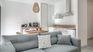 Un saló modern amb un sofà gris, una taula de dinar amb elements per esmorzar, i una cuina amb armaris blancs i una campana extractora d'acer inoxidable.