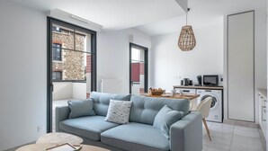 Un saló modern amb un sofà gris, una taula de fusta petita i una zona de cuina amb electrodomèstics.