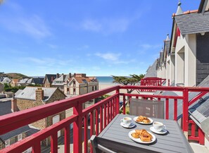 A balcony with a table set for breakfast, overlooking a coastal town with traditional architecture and a clear sky.