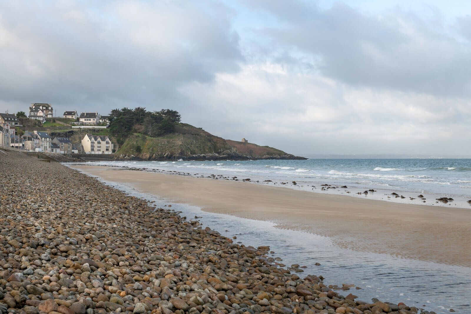 A pebble beach with a rocky shoreline, a small hill with buildings, and the ocean in the background.