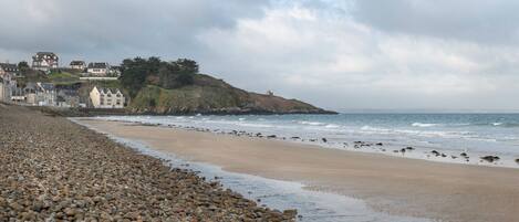 A pebble beach with a rocky shoreline, a small hill with buildings, and the ocean in the background.