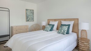 Un dormitorio ordenado con una cama grande, una mesita de noche con lámpara, un revistero de mimbre y una obra de arte colgada en la pared.