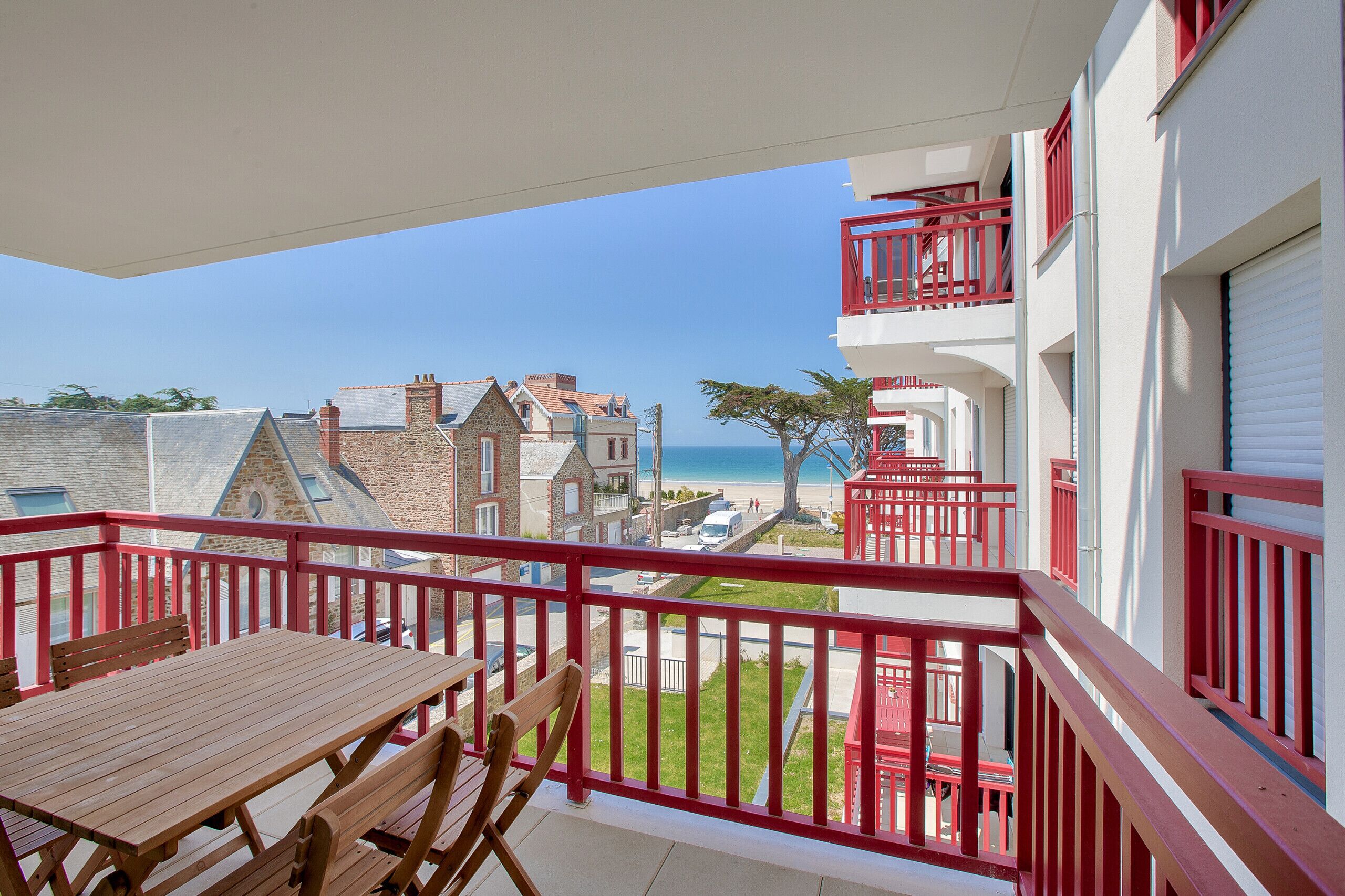 A balcony with a wooden table and chairs, overlooking a beach and buildings.