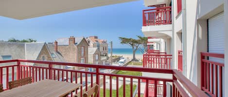 A balcony with a wooden table and chairs, overlooking a beach and buildings.