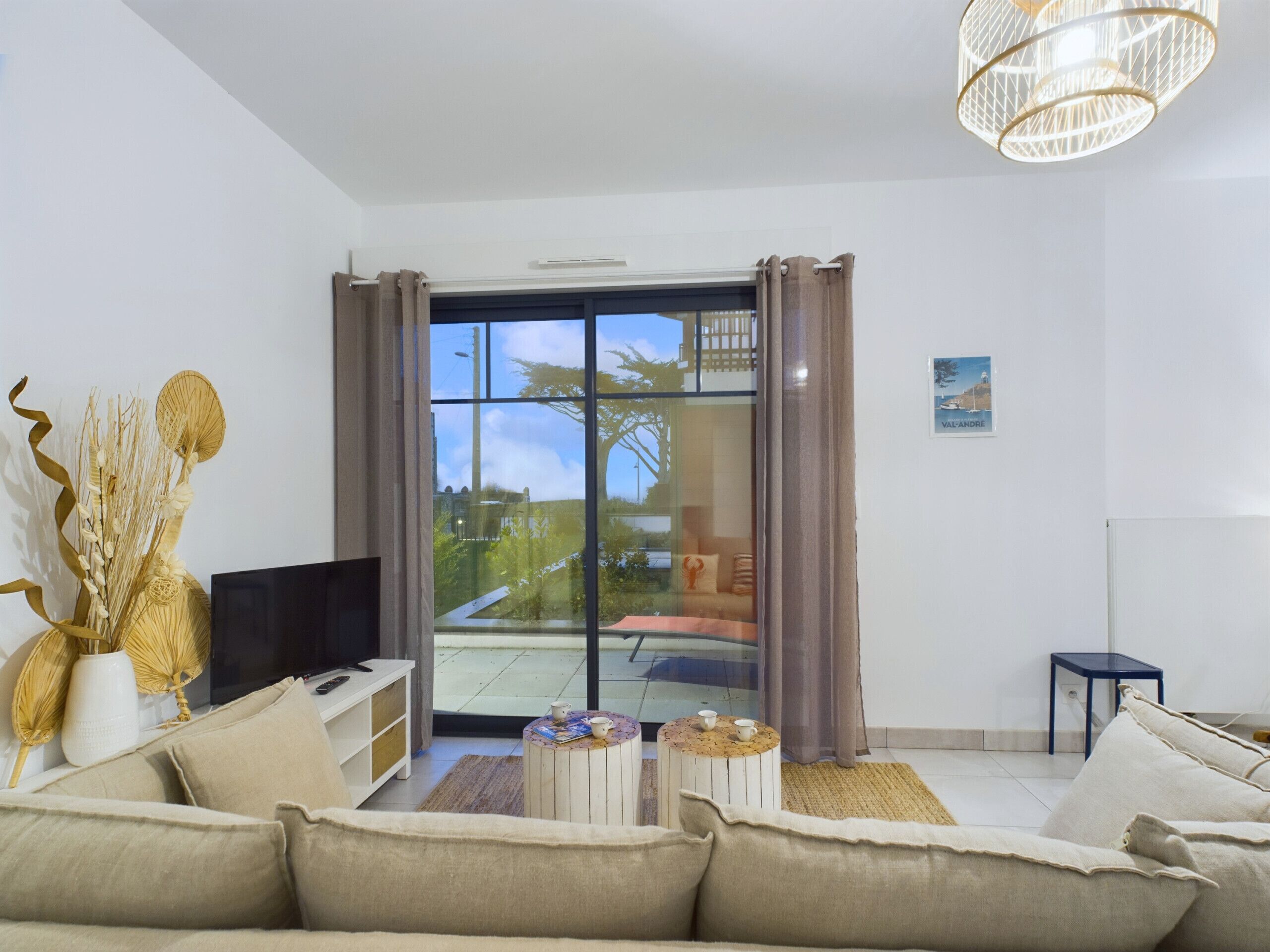 A modern living room with a sofa, coffee table, and a TV. There's a large window with curtains, a chandelier, and a small table with cups.