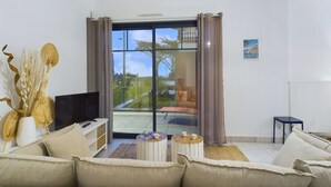 Een moderne woonkamer met een bank, salontafel en een televisie. Er is een groot raam met gordijnen, een kroonluchter en een klein tafeltje met kopjes.