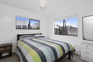 Een slaapkamer met een bed, nachtkastje, dressoir en twee ramen.