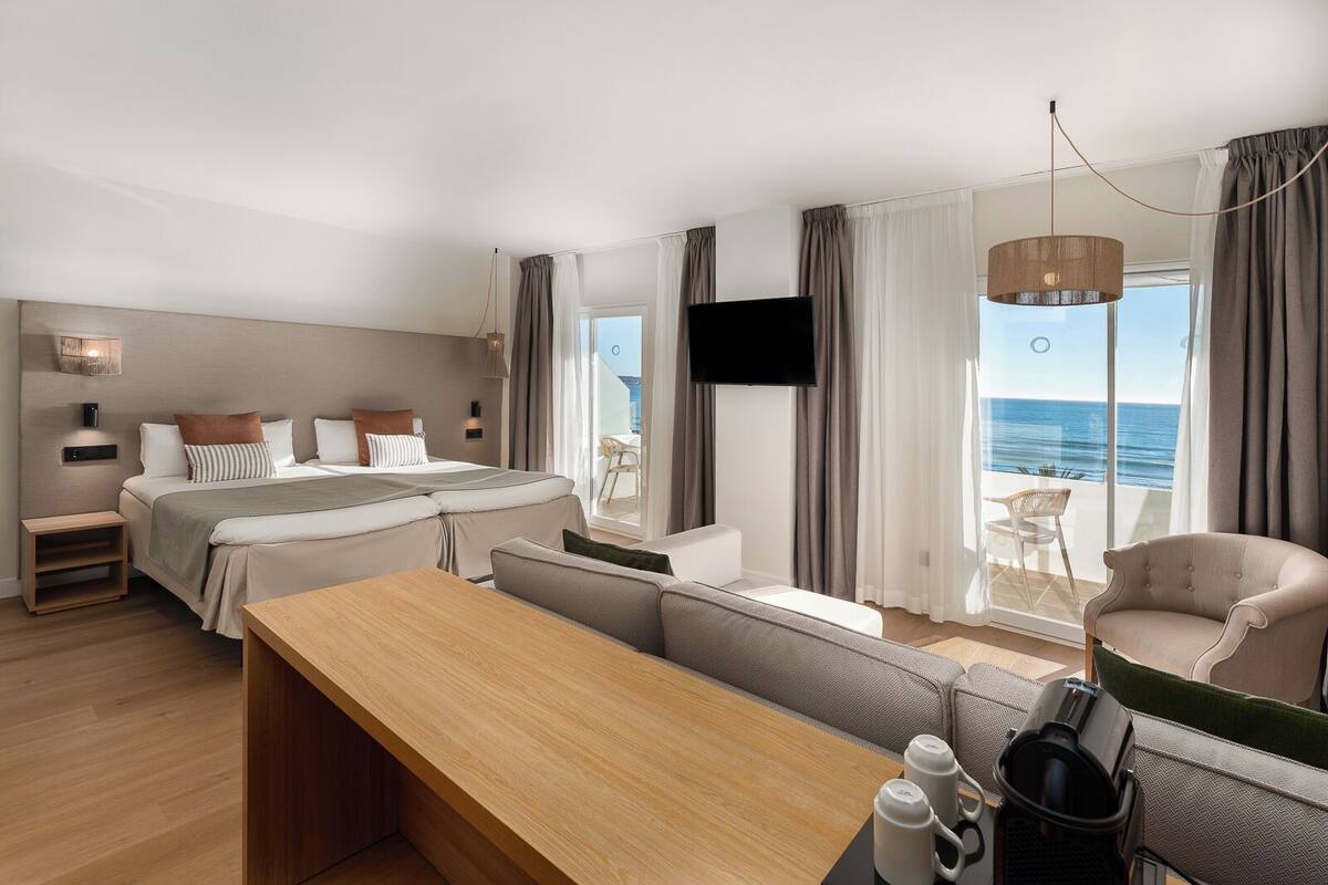 A modern hotel room with a bed, a sofa, a dining table, and a TV.