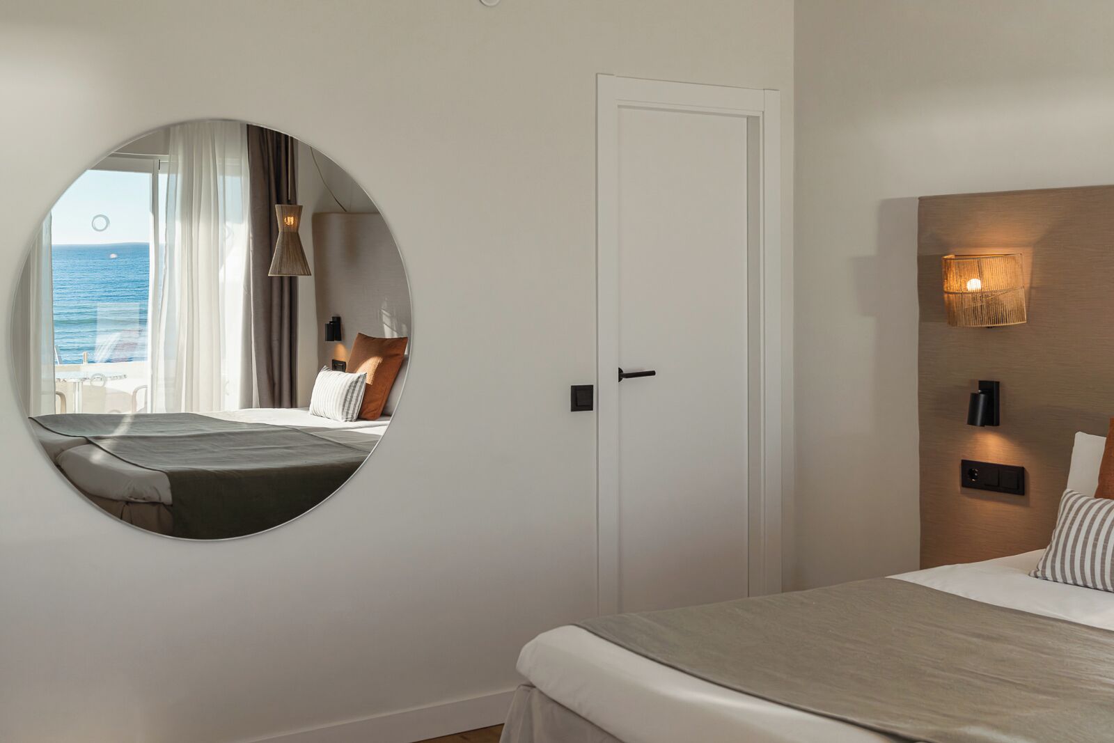 A hotel room with a circular mirror, a bed with a wooden headboard, a bedside table, and a view of the sea through a window.
