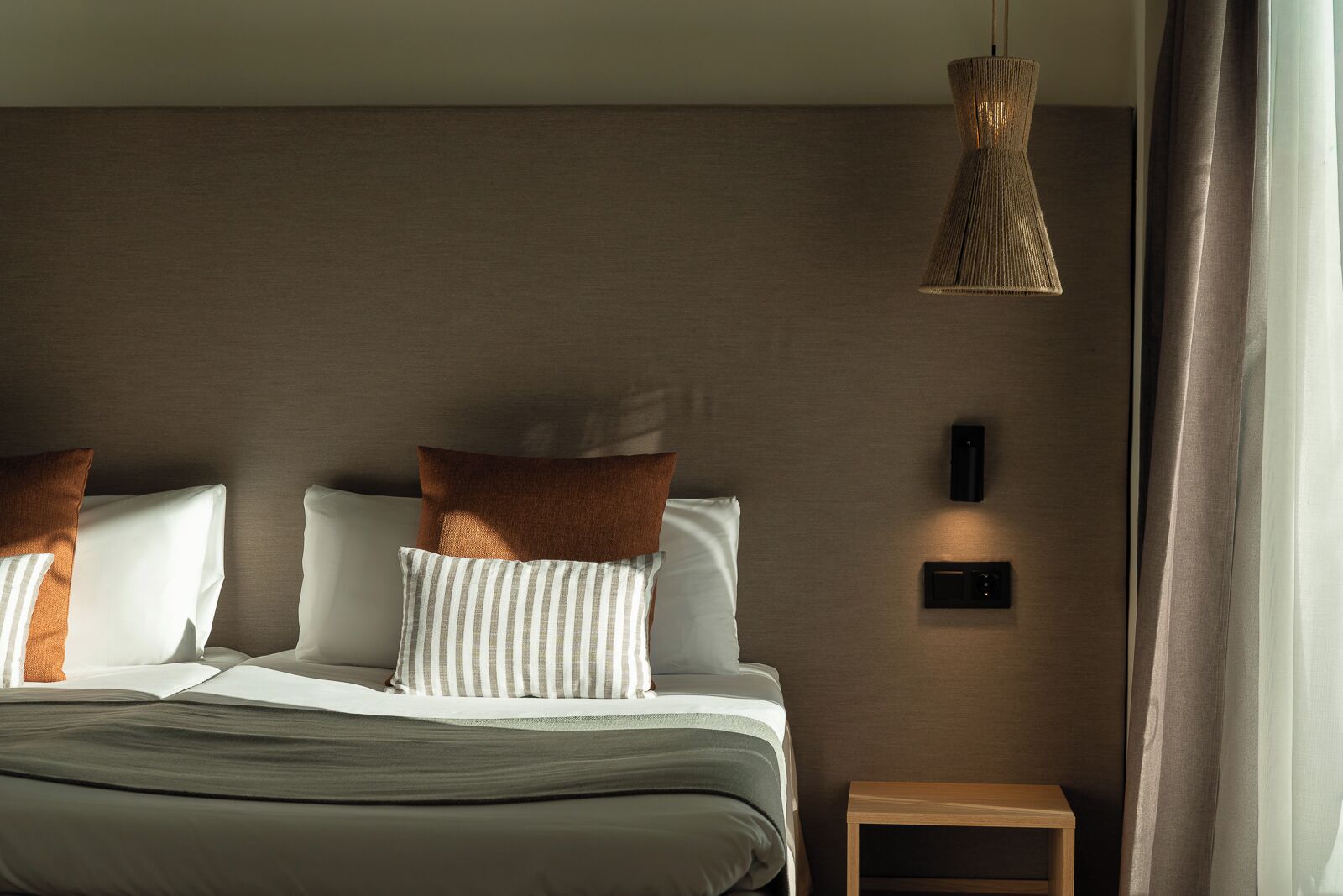 A modern hotel room with a bed, pillows, a bedside table, a wall-mounted light, and a window with curtains.