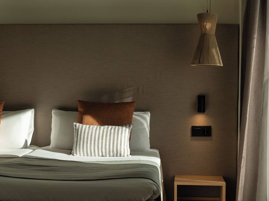 A modern hotel room with a bed, pillows, a bedside table, a wall-mounted light, and a window with curtains.