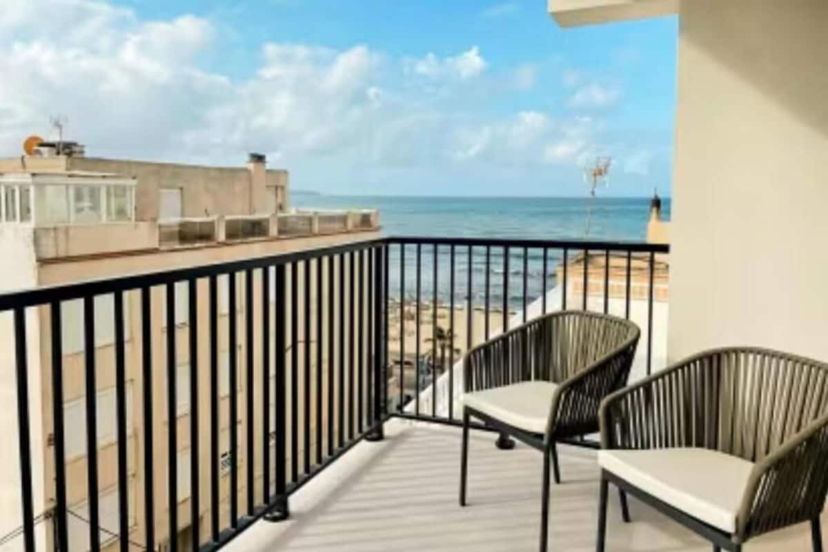 A balcony with two wicker chairs, a view of the sea, and a building in the background.