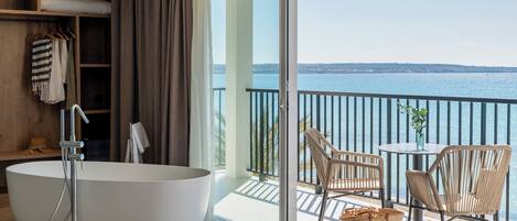 Un baño moderno con una bañera independiente, ducha y un balcón con cerramiento de vidrio, mobiliario de mimbre y vista al mar.