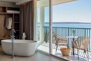 Un baño moderno con una bañera independiente, ducha y un balcón con cerramiento de vidrio, mobiliario de mimbre y vista al mar.