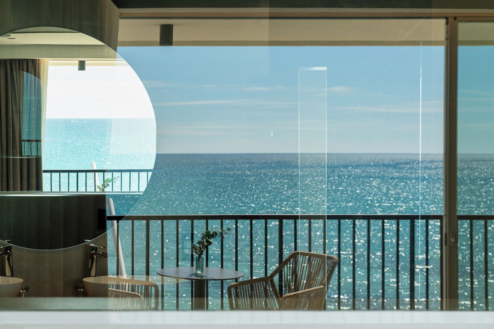 A balcony with a view of the sea, a round table, two wicker chairs, and a vase with flowers.