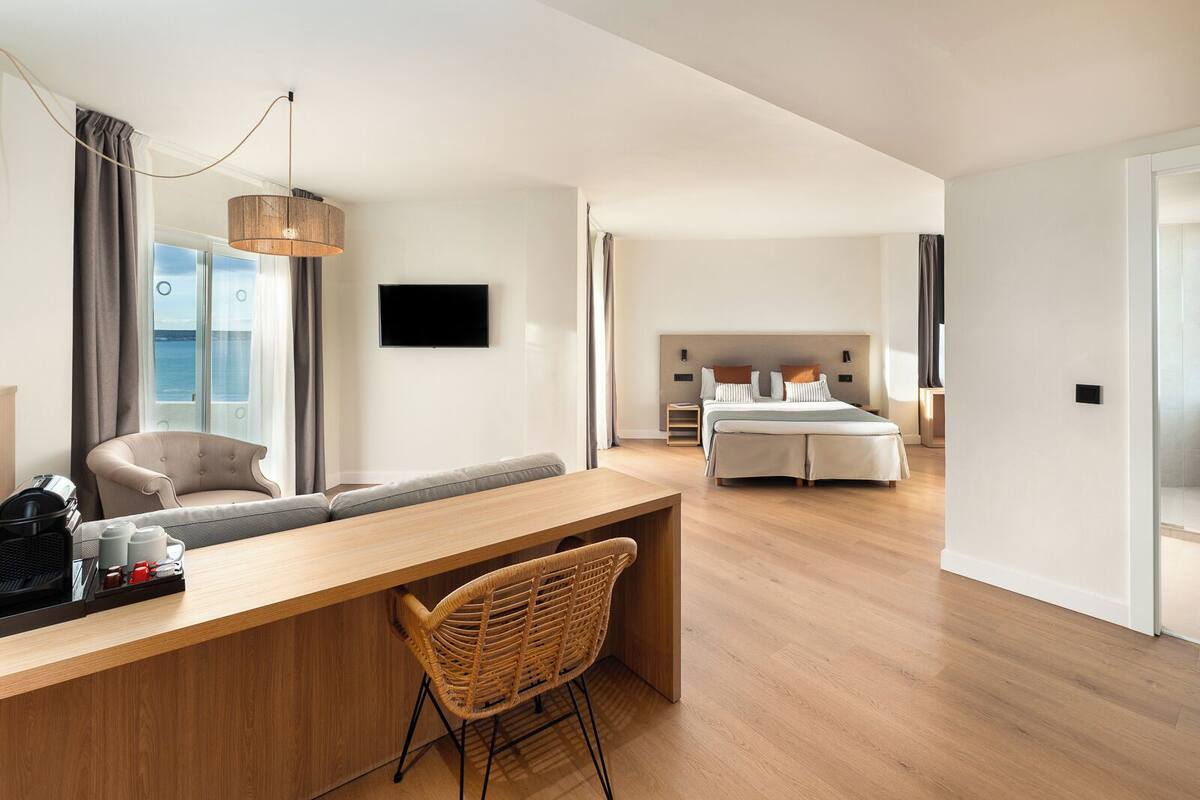 A modern hotel room with a wooden desk, wicker chair, a large bed, and a TV.