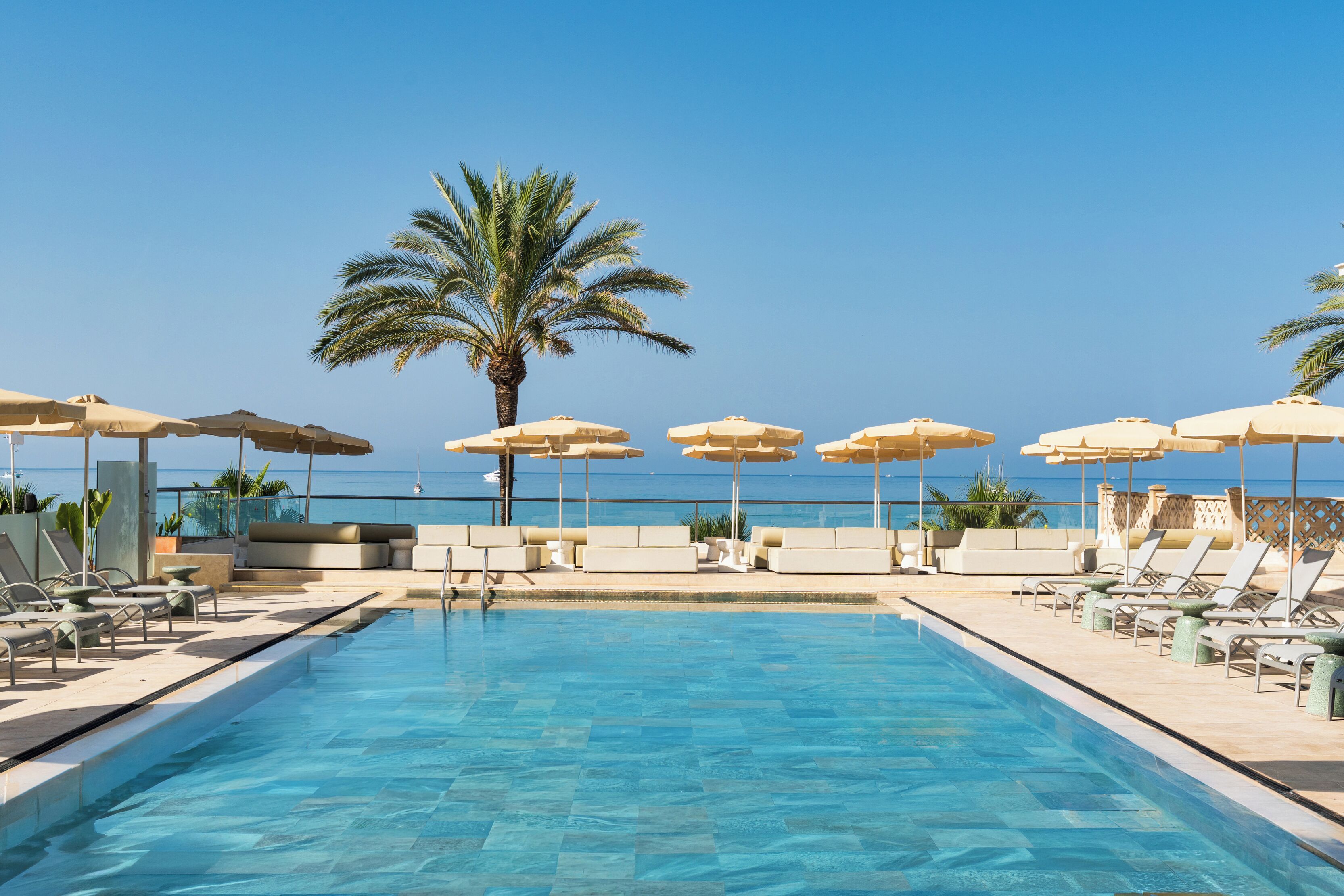 Une piscine entourée de palmiers, avec des chaises longues et des parasols, offrant une vue sur la mer.