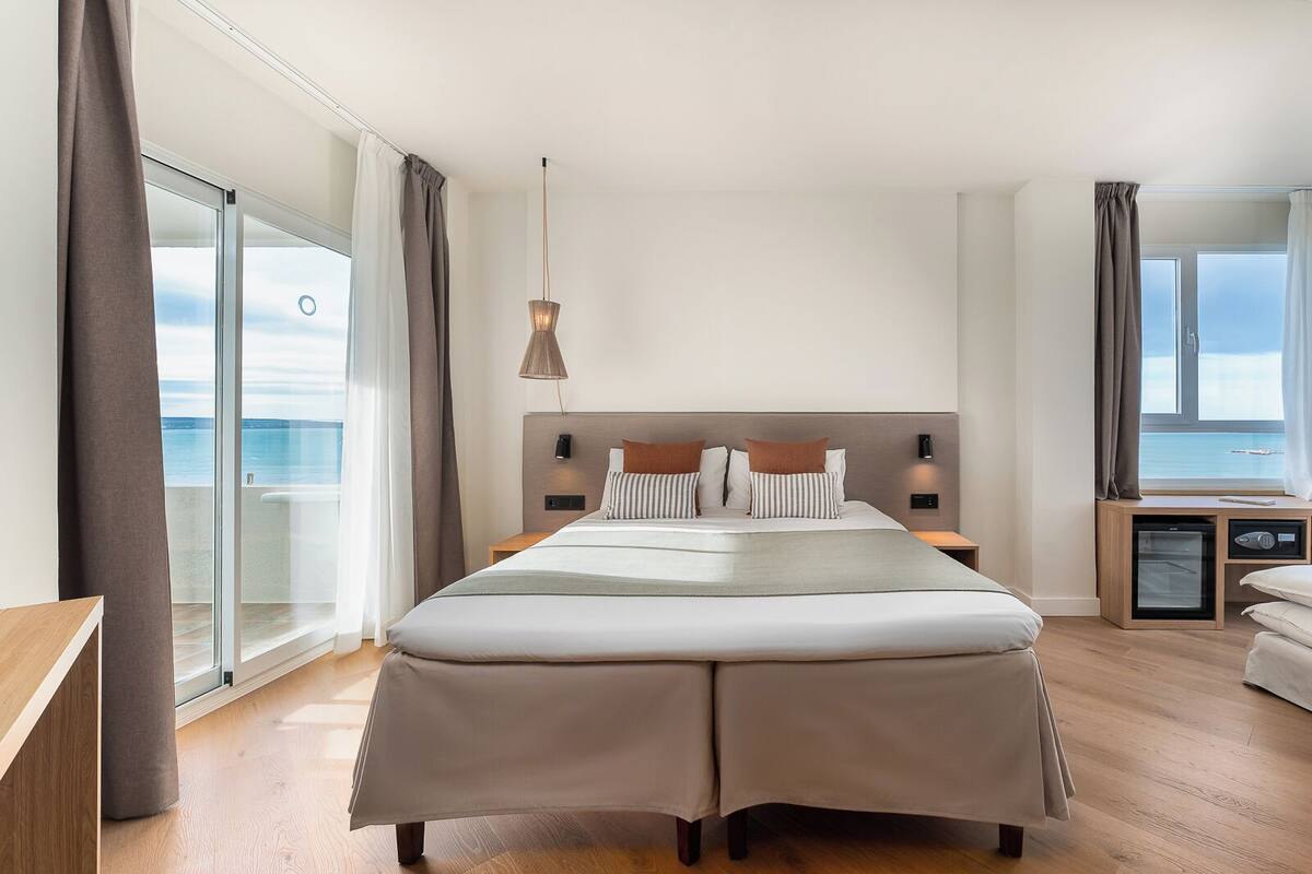 A modern hotel room with a large bed, a view of the sea, and a small kitchenette.