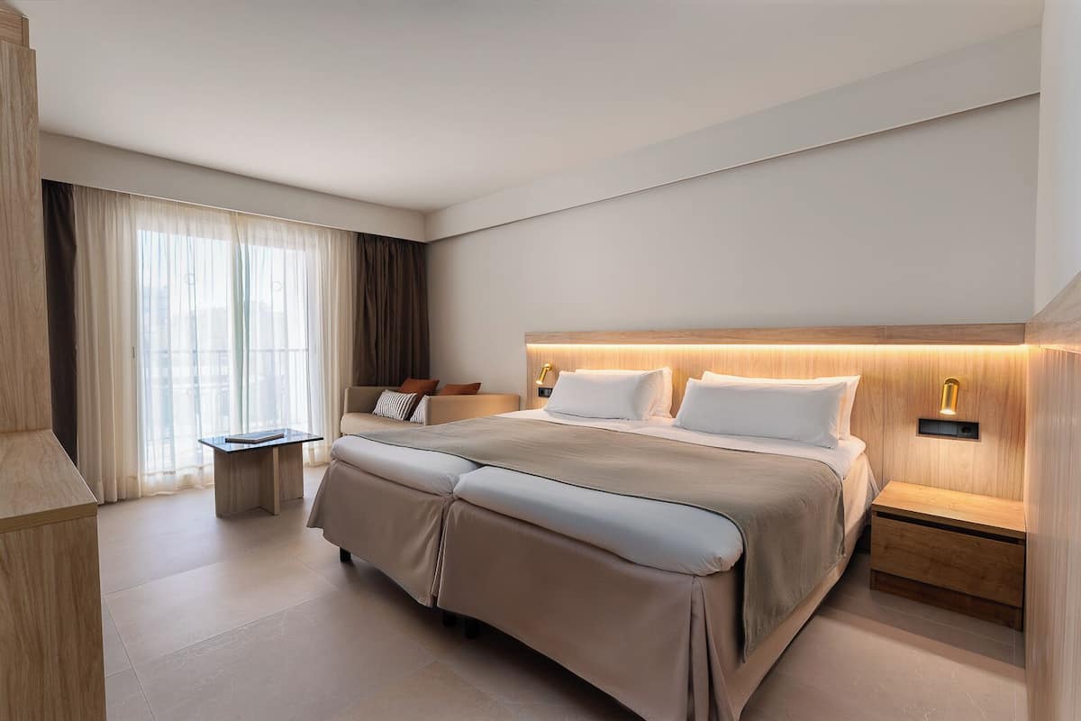 A modern hotel room with a large bed, bedside tables, a desk, and a view of the city through large windows.