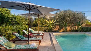 Un área junto a la piscina con sillones de descanso y sombrillas, rodeada de exuberante vegetación.