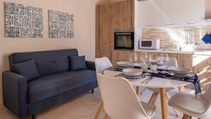 Una cocina moderna con armarios blancos, un microondas y una encimera de madera. Hay una mesa de comedor preparada con platos, vasos y cubiertos. Un sofá azul se encuentra frente a la cocina.
