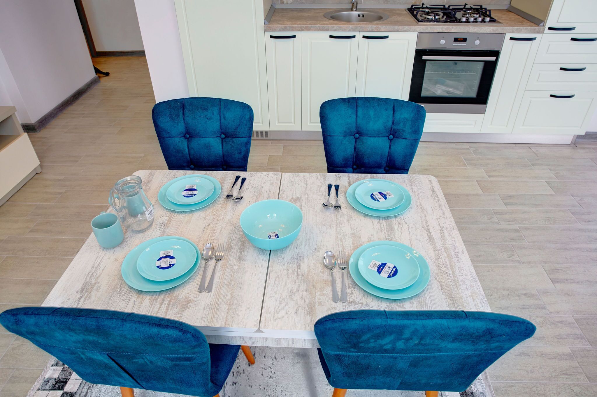 Una mesa de comedor dispuesta con platos, tazones y cubiertos azules, rodeada de sillas azules a juego en una cocina moderna con armarios blancos y una estufa a gas.