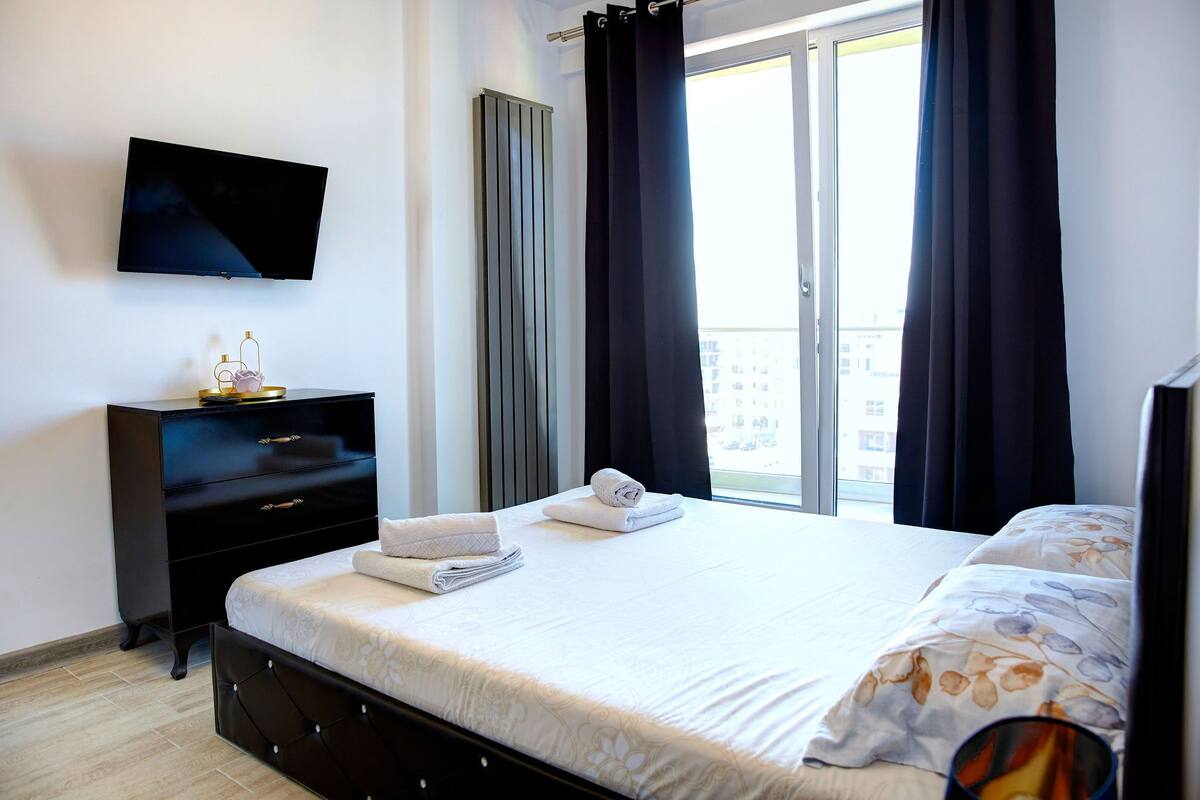 Una habitación de hotel con cama, televisor, cómoda y ventana con cortinas.