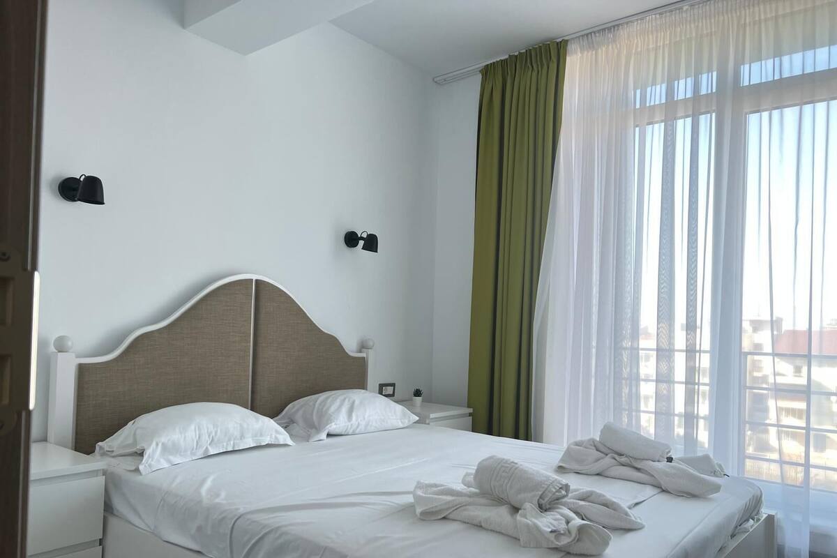 Una cama bien hecha con ropa de cama y toallas blancas, una mesita de noche con lámpara, y vistas a edificios a través de cortinas traslúcidas.