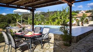 An outdoor patio with a table set for dining, a pool, and lush greenery in the background. - 1841 Petrino (Kefalonia)