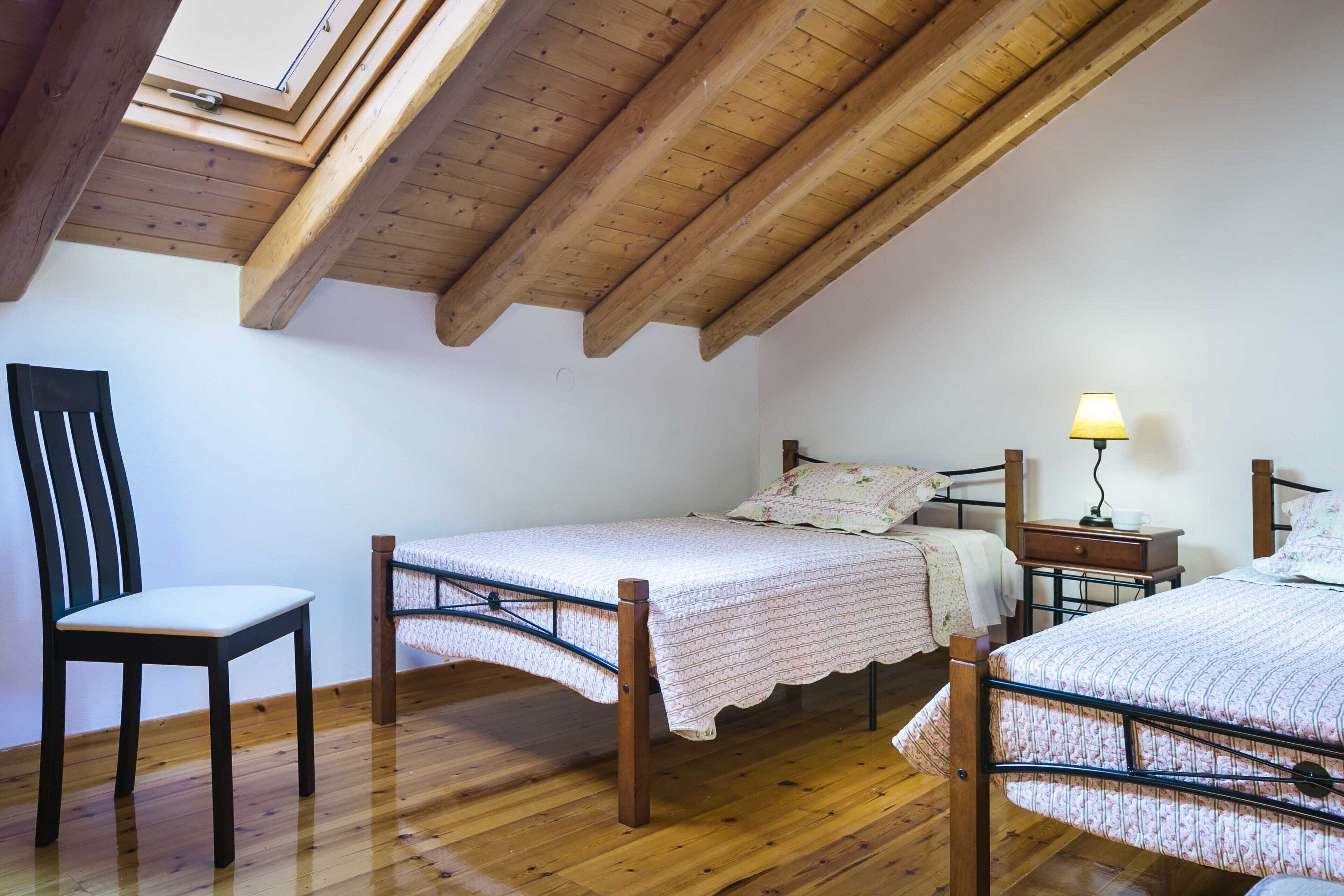 A bedroom with a sloped ceiling, wooden flooring, two beds, a chair, a nightstand, and a lamp.