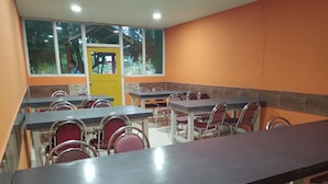 Comedor con varias mesas y sillas, una puerta amarilla y ventanas con vistas a follaje.