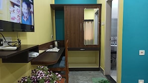 Una habitación pequeña y luminosa con un escritorio de madera, un televisor, un espejo y una pequeña mesa con flores.