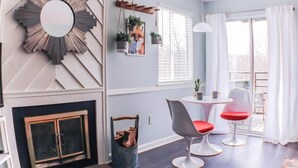 Un salon moderne avec une cheminée, un miroir rond, un coin repas avec deux chaises et une fenêtre avec des stores.