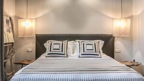A neatly made bed with a striped bedspread and two square pillows, flanked by two bedside tables with lamps, in a room with a plain wall and a painting on the left. - Avithos Resort (Kefalonia)
