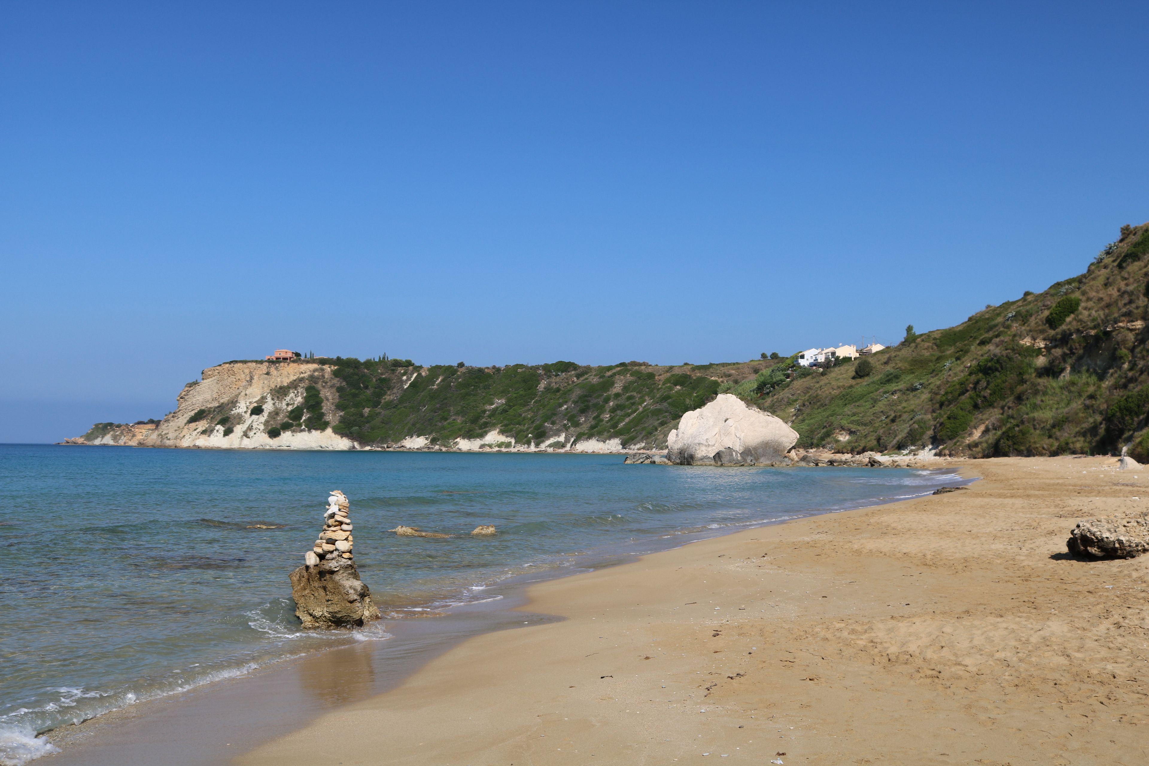 A beach with clear water, a rocky outcrop, and a hill with buildings.
