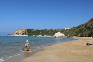 Una playa con agua cristalina, un promontorio rocoso y una colina con edificios.