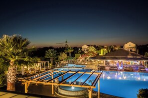A well-lit outdoor swimming pool area with lounge chairs and a bar, surrounded by palm trees and a building with a sloped roof. - Avithos Resort (Kefalonia)