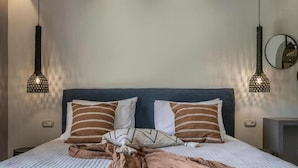 A neatly made bed with striped pillows and a blanket, two pendant lights, and a round mirror on the wall. - Avithos Resort (Kefalonia)