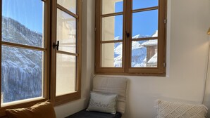 Ein Zimmer mit einer Bank und einem kleinen Sofa, beide mit Kissen. Zwei Fenster mit Holzrahmen bieten Blick auf verschneite Berge.