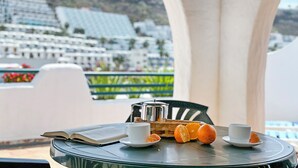 A table with a book, two cups, and oranges, overlooking a coastal town with buildings and mountains. - Laguna Apartments (Mogan)