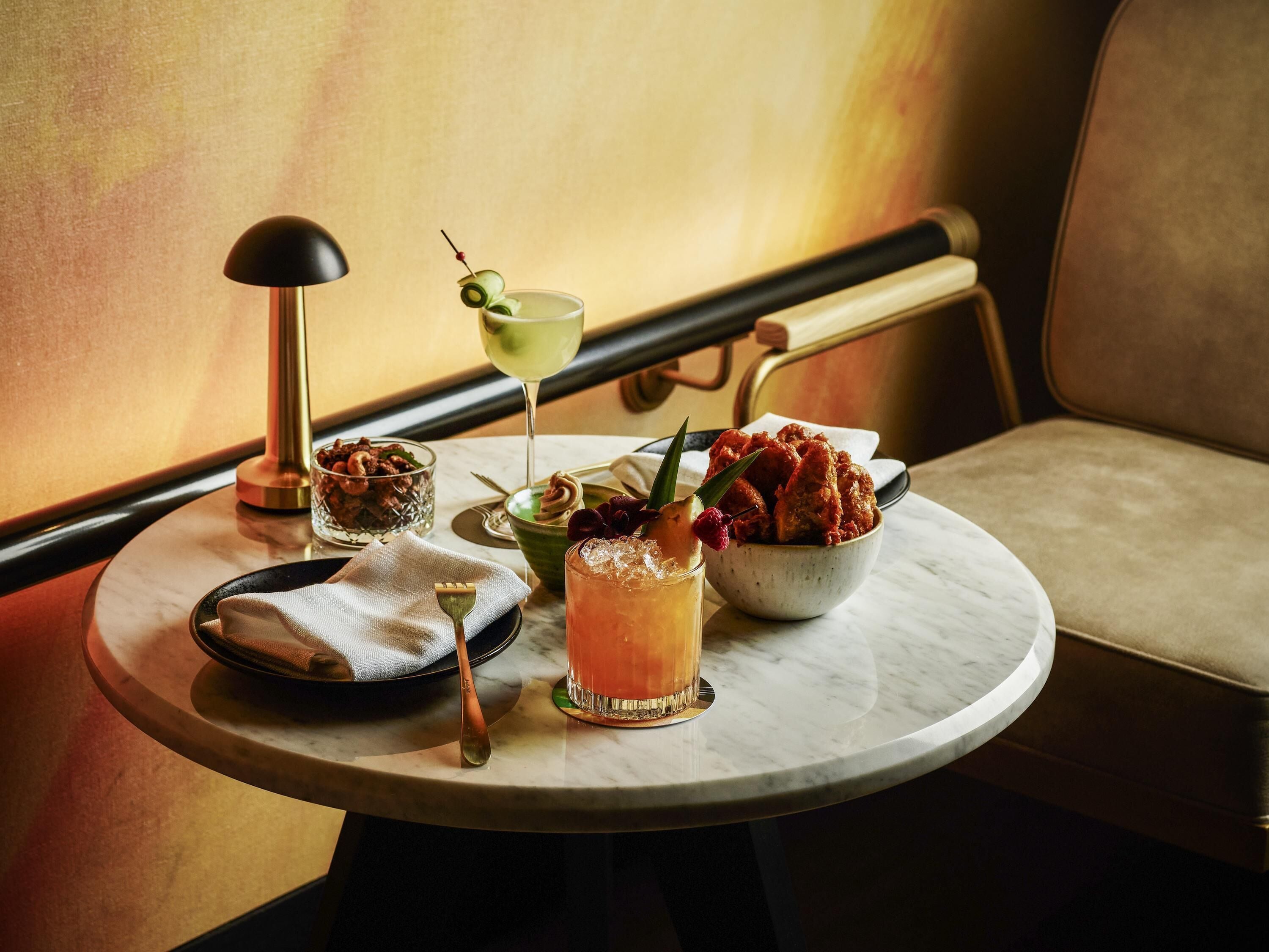 Una mesa redonda con una copa de martini, un bol de pollo frito, un cuenco pequeño de nueces, una cuchara y una servilleta.