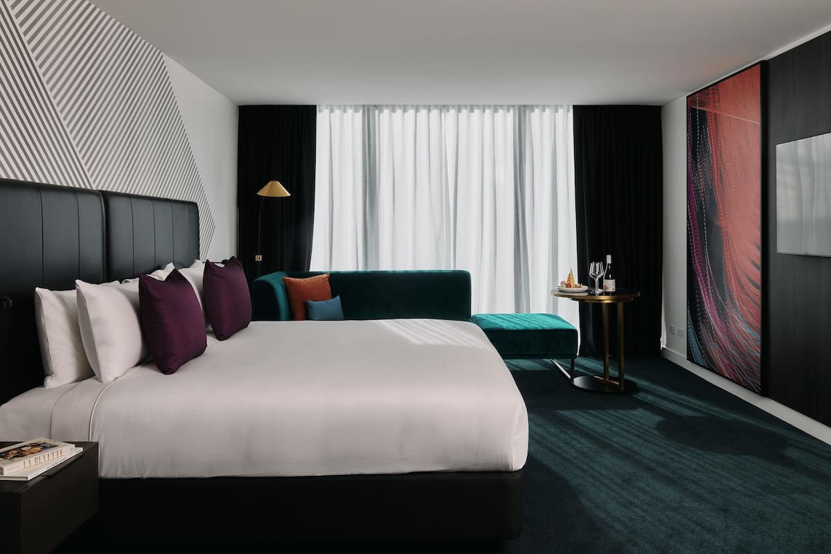 Una habitación de hotel moderna con una cama grande, un sofá, una mesita con una lámpara y un cuadro abstracto de gran tamaño en la pared.