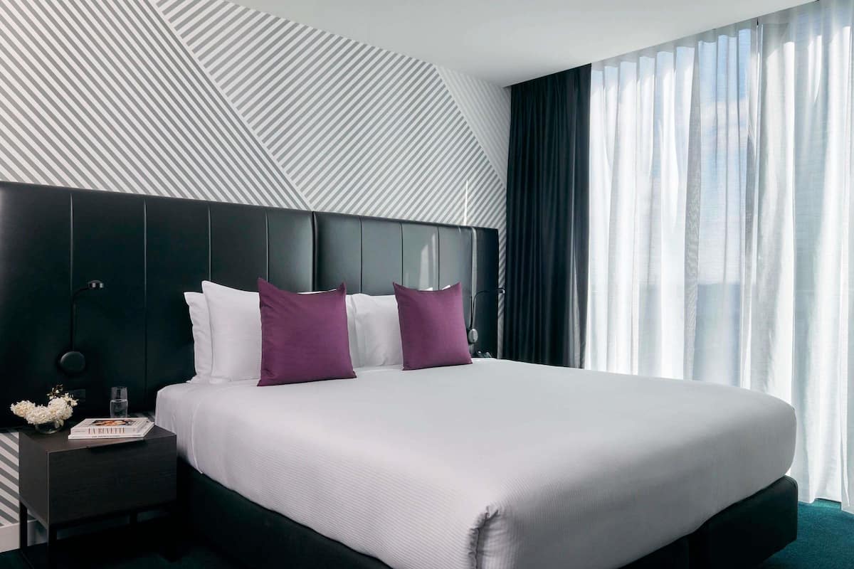 Habitación de hotel moderna con una cama grande, cabecera oscura y vistas al cielo a través de cortinas translúcidas.