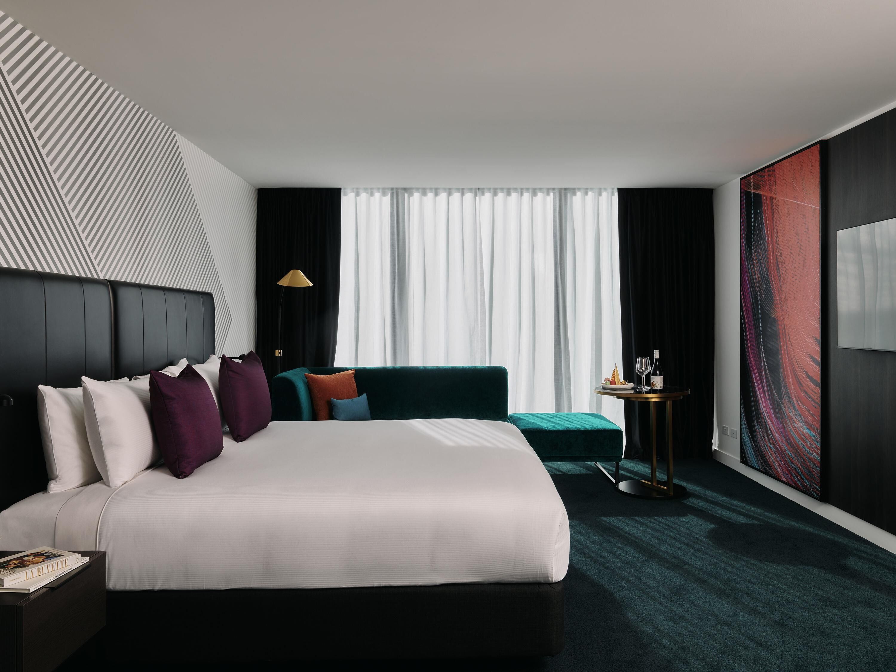 Quarto de hotel moderno com uma cama grande, um sofá, uma mesa lateral com abajur e uma janela ampla com cortinas.