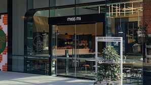 Un edificio moderno con fachada de vidrio, un letrero que dice 'miss mi' y mesas al aire libre visibles a través de las ventanas.